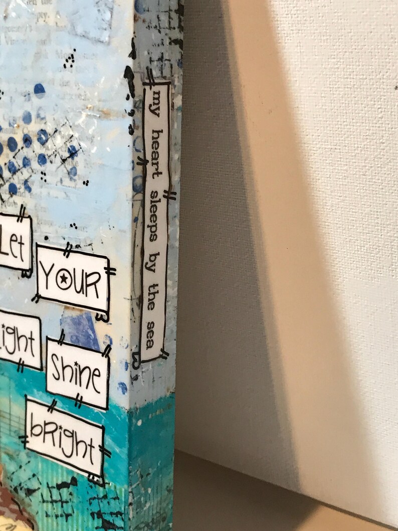 May include: A blue and white canvas painting with the text "Let your light shine bright" and "my heart sleeps by the sea" on the side.