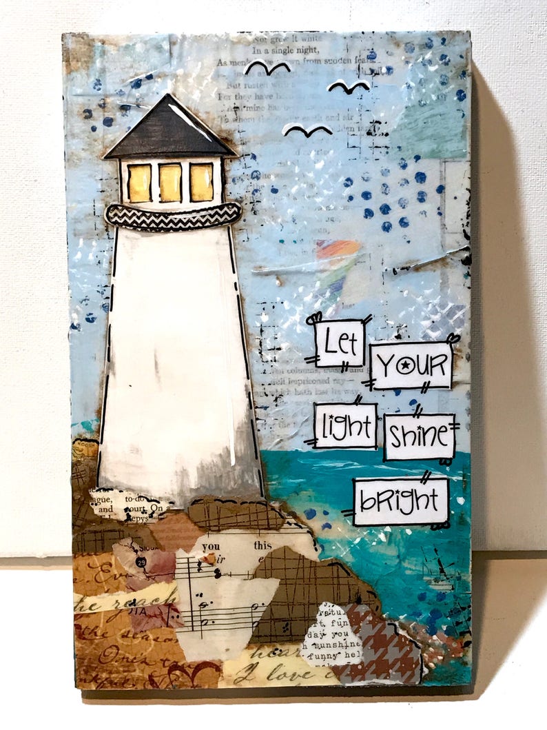 May include: A mixed media art piece on a rectangular board featuring a white lighthouse with a black roof and yellow windows. The background includes collage elements of text and blue paint. The text "Let YOUR light shine bRight" is displayed.