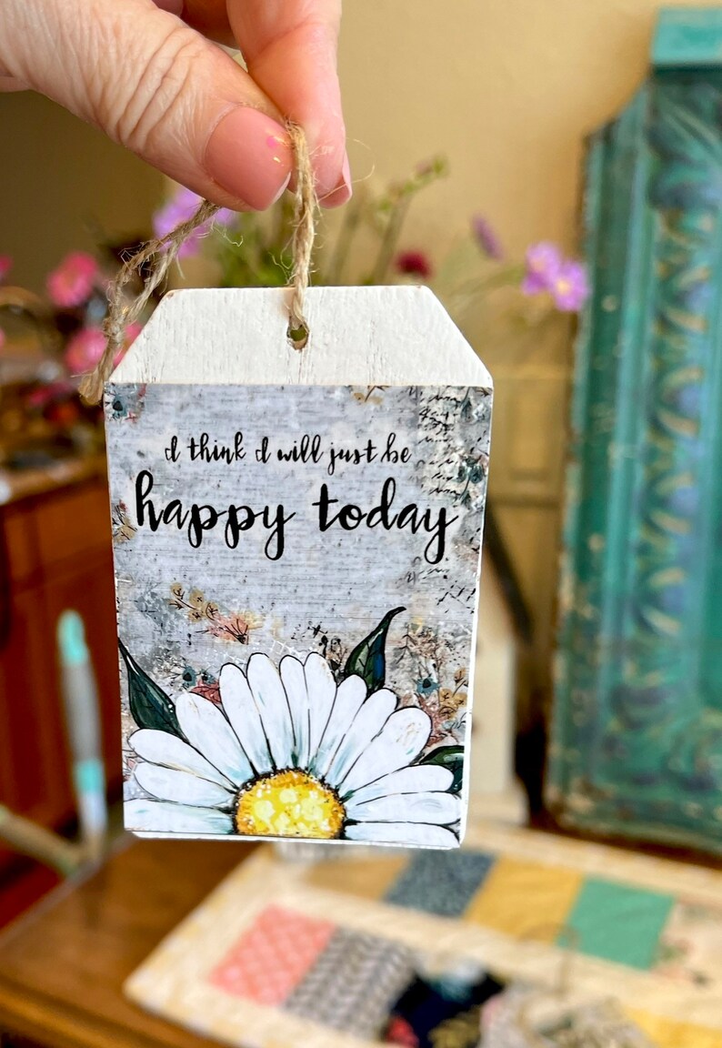May include: A wooden tag with a jute string for hanging. The tag features the text "I think I will just be happy today" above a large white daisy with a yellow center. The background has a distressed, floral design.