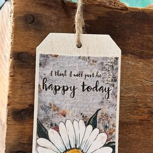 Puede incluir: Una etiqueta de madera con el texto "I think I will just be happy today" en escritura negra. La etiqueta presenta una margarita de estilo acuarela en la parte inferior y est&aacute; suspendida por un cordel natural. El fondo tiene un aspecto r&uacute;stico y desgastado.
