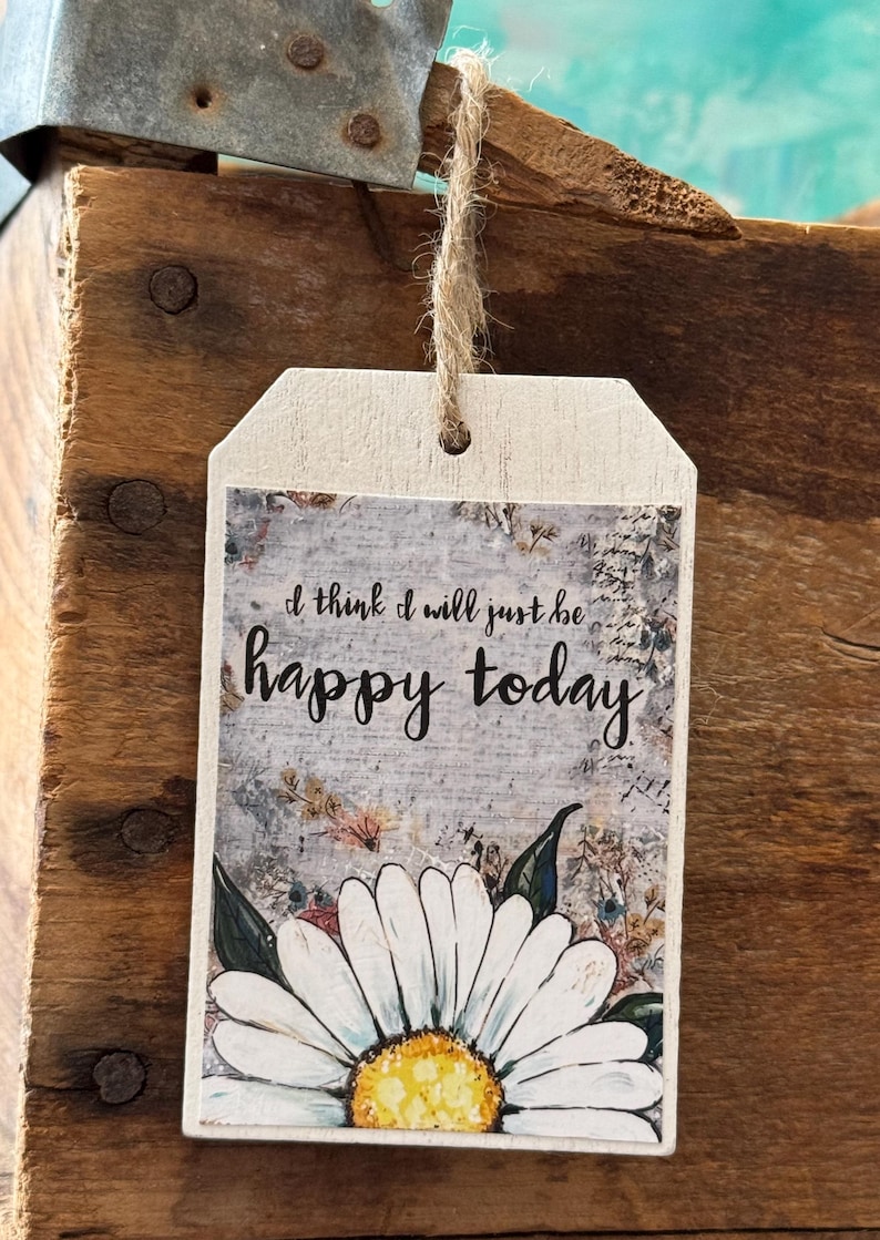 Puede incluir: Una etiqueta de madera con el texto "I think I will just be happy today" en escritura negra. La etiqueta presenta una margarita de estilo acuarela en la parte inferior y est&aacute; suspendida por un cordel natural. El fondo tiene un aspecto r&uacute;stico y desgastado.
