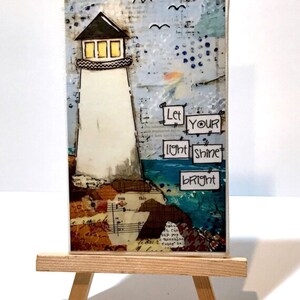 May include: A rectangular mixed media art piece featuring a white lighthouse with yellow windows, set against a collage background of text and blue tones. The text "Let YOUR light shine bRight" is displayed in black ink on white paper squares.