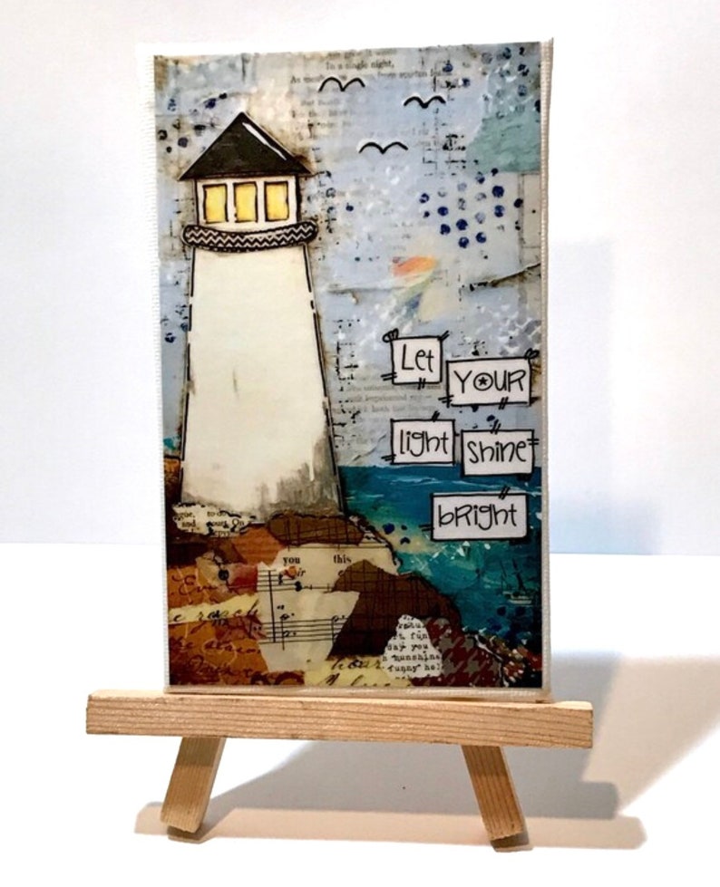 May include: A rectangular mixed media art piece featuring a white lighthouse with yellow windows, set against a collage background of text and blue tones. The text "Let YOUR light shine bRight" is displayed in black ink on white paper squares.