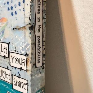 May include: A canvas art piece featuring a blue and white abstract background with collage-style text boxes. The side edge displays the text "believe in the possibilities," while the front shows "Let YOUR light SHINE bright" in black ink on white paper.