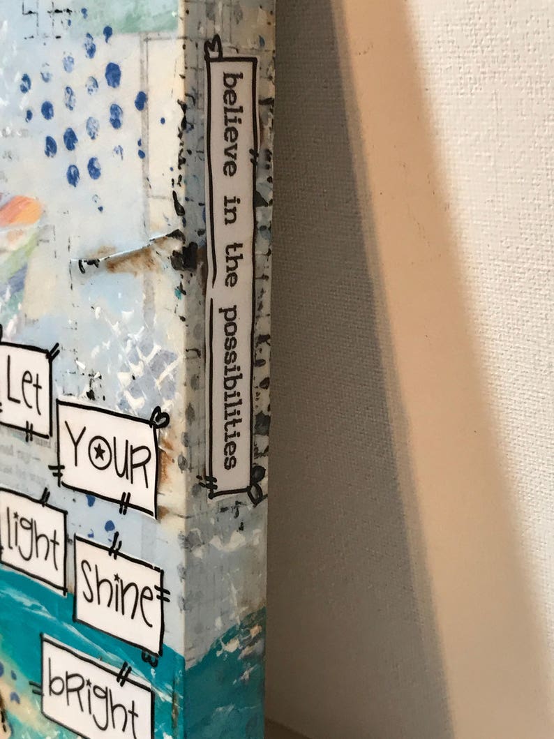 May include: A canvas art piece featuring a blue and white abstract background with collage-style text boxes. The side edge displays the text "believe in the possibilities," while the front shows "Let YOUR light SHINE bright" in black ink on white paper.