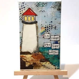 May include: A mixed media painting on canvas featuring a lighthouse with a red roof and a white tower. The lighthouse is on a cliff with brown and blue textured details. The painting has a blue background with white birds and the text "Let your light shine bright".