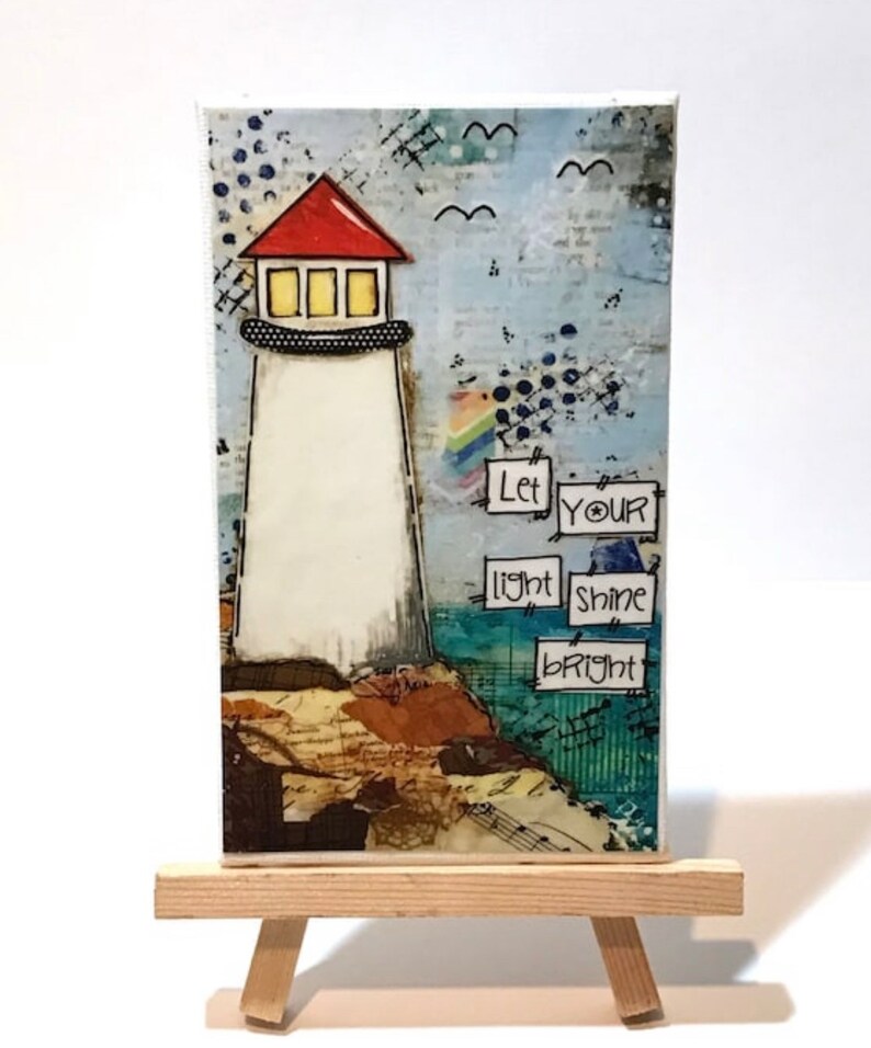May include: A mixed media painting on canvas featuring a lighthouse with a red roof and a white tower. The lighthouse is on a cliff with brown and blue textured details. The painting has a blue background with white birds and the text "Let your light shine bright".