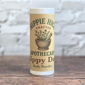 May include: A cylindrical cardboard container of body powder with a tan label. The label features green text reading Hippie Hick, the word Crafted, an illustration of a mortar and pestle with herbs, and the text Apothecary Hippy Dust Body Powder.