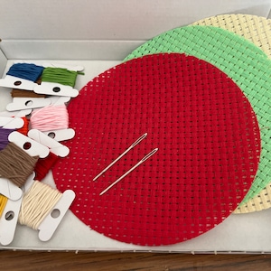 Puede incluir: Un conjunto de tres aros de bordado redondos en rojo, verde y crema, con una variedad de hilos de bordar de colores y dos agujas en una caja blanca.