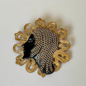 May include: A gold-toned brooch featuring a black silhouette of a woman's head with braided hair. The hair is made of a chain-like material and the brooch has a decorative, floral-shaped design.