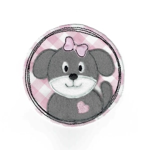 May include: A round, gray dog applique with a pink bow and heart. The dog is centered on a pink and white gingham background, framed by a black stitched border. The applique is likely for a craft project or clothing embellishment.