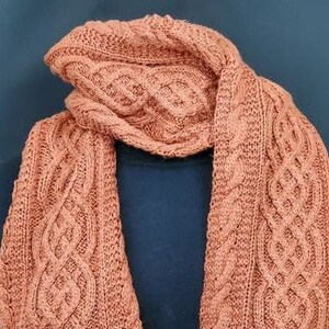 May include: A long, knitted scarf in a warm terracotta color with a cable knit pattern. The scarf is folded in half and draped over a dark surface.