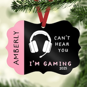 May include: A black and pink Christmas ornament with the name "AMBERLY" in pink. The ornament features a white headset graphic and the text "CAN'T HEAR YOU I'M GAMING 2025". A red ribbon is attached for hanging.