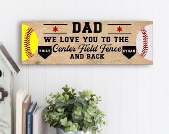 May include: Wooden sign with a softball and baseball design. The sign reads "DAD WE LOVE YOU TO THE CENTER FIELD FENCE AND BACK" with the names "EMILY" and "ETHAN" below. The sign is on a white wall.