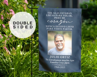 May include: A blue and white memorial garden flag with a photo of a man and the text "Tus alas estaban listas para volar, pero mi corazón nunca estuvo listo para verte partir. Julio Ortiz Te extrañamos mucho Apa Familia Cruz Jijon 1960-2023"