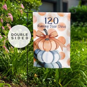 May include: A double-sided garden flag featuring a stack of pumpkins in shades of orange, blue, and white, topped with a brown bow. The flag displays the text "120 Running Deer Drive" against a backdrop of orange polka dots. The flag is displayed on a black metal stand.