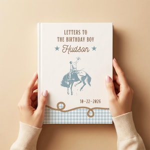 May include: A white hardcover book titled Letters to the Birthday Boy Hudson with a blue illustration of a cowboy riding a bucking horse. The cover features two blue stars, the date 10-22-2026, and a brown rope graphic over a light blue gingham pattern.