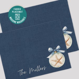 May include: Two navy blue fabric placemats with a linen texture. Each placemat features a watercolor design of a starfish inside a glass ornament with a blue bow. The text "The Millers" is printed on the bottom left of the placemats. The placemats are machine washable.