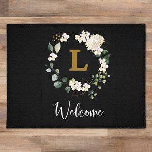 May include: Black doormat with a floral wreath design featuring white flowers and green leaves. The wreath surrounds a gold letter "L" and the word "Welcome" is printed below the wreath.