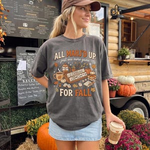 May include: A charcoal gray short-sleeve t-shirt featuring a graphic print with the text "ALL MAHJ'D UP just one more game FOR FALL" surrounded by illustrations of mahjong tiles, a coffee mug, a French press, pumpkins, and autumn leaves.