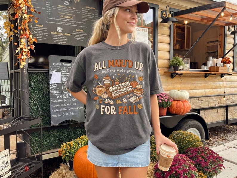 May include: A charcoal gray short-sleeve t-shirt featuring a graphic print with the text "ALL MAHJ'D UP just one more game FOR FALL" surrounded by illustrations of mahjong tiles, a coffee mug, a French press, pumpkins, and autumn leaves.