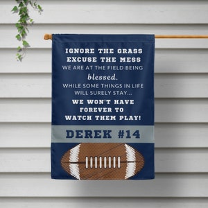May include: A blue and gray garden flag with a football design and the text "IGNORE THE GRASS EXCUSE THE MESS WE ARE AT THE FIELD BEING blessed. WHILE SOME THINGS IN LIFE WILL SURELY STAY... WE WON'T HAVE FOREVER TO WATCH THEM PLAY! DEREK #14"