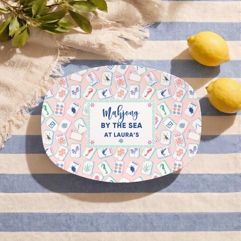 May include: Oval-shaped pink serving platter with a Mahjong tile pattern and the text "Mahjong by the Sea at Laura's." The platter is surrounded by lemons and a blue and white striped tablecloth.