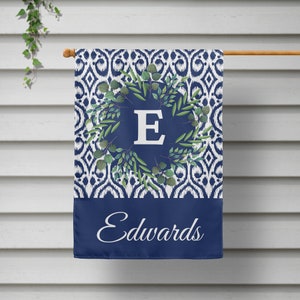 May include: A blue and white patterned garden flag with a green wreath and the letter "E" in white. The bottom of the flag is solid blue with the name "Edwards" in white cursive.
