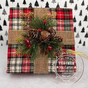 K&ouml;nnte beinhalten: Eine Weihnachtsgeschenkbox, verpackt in rot-schwarz kariertem Stoff, mit Juteband und festlichem Gr&uuml;n, Tannenzapfen und roten Beeren. Ein klarer Ornament mit goldenem Band liest sich "First Year as a Teacher Ms. Carter Windsong Ranch Elementary 2023-2024."