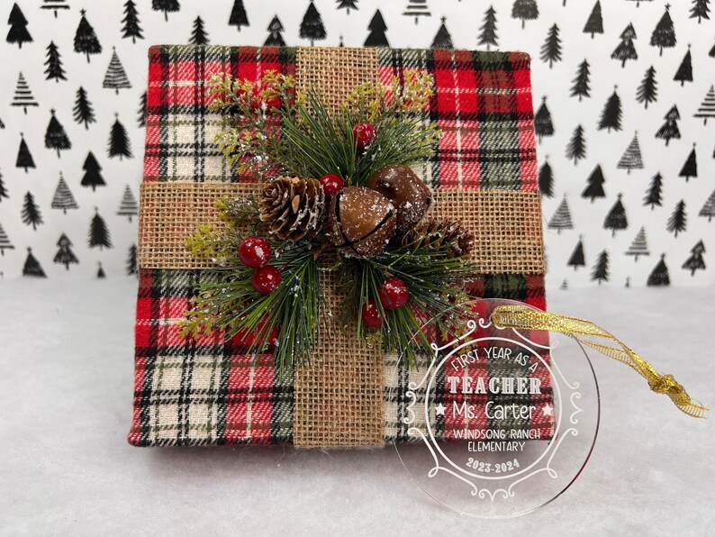K&ouml;nnte beinhalten: Eine Weihnachtsgeschenkbox, verpackt in rot-schwarz kariertem Stoff, mit Juteband und festlichem Gr&uuml;n, Tannenzapfen und roten Beeren. Ein klarer Ornament mit goldenem Band liest sich "First Year as a Teacher Ms. Carter Windsong Ranch Elementary 2023-2024."