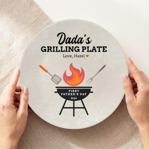 May include: A round, off-white plate with the text "Dada's GRILLING PLATE" and "Love, Hazel". The plate features a grill graphic with a flame, spatula, and fork. The text "FIRST FATHER'S DAY 2026" is also included.