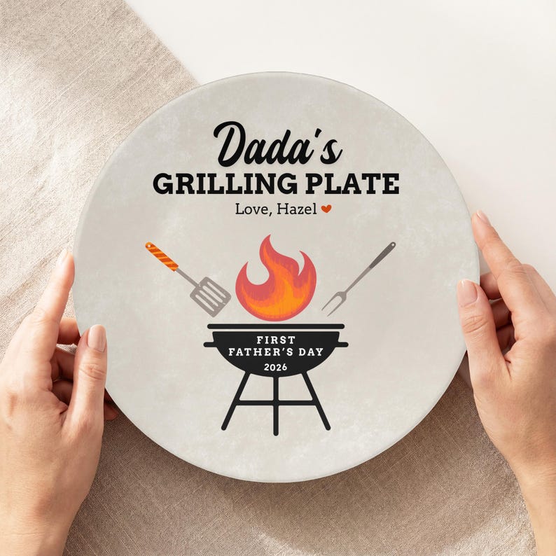 May include: A round, off-white plate with the text "Dada's GRILLING PLATE" and "Love, Hazel". The plate features a grill graphic with a flame, spatula, and fork. The text "FIRST FATHER'S DAY 2026" is also included.
