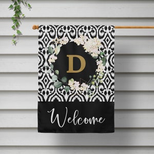 May include: Black and white welcome garden flag with a floral wreath and the letter D in gold. The flag has a black background with a white welcome message.