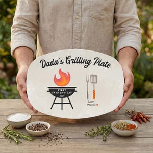 May include: An oval grilling plate with the text "Dada's Grilling Plate" and a grill graphic. The plate also features a flame, a spatula, and the text "First Father's Day 2026". The plate is held by a person.