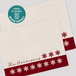 May include: Two cream-colored fabric placemats with a red border featuring white snowflake designs. The text "The Harrisons" is printed in a cursive font. A teal circle indicates the placemats are machine washable.