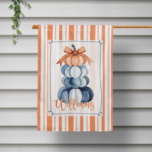 May include: A decorative garden flag featuring a stack of pumpkins in shades of orange, blue, and white, topped with an orange bow. The flag has a striped background in orange and white, with the name "Williams" in script.