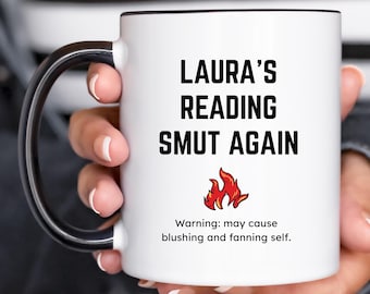 May include: White ceramic mug with a black handle and rim. The mug features the text "LAURA'S READING SMUT AGAIN" in black, with a small flame graphic and the warning: "may cause blushing and fanning self."