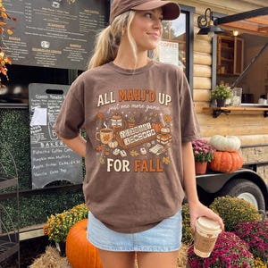 May include: A brown short-sleeve t-shirt featuring a graphic print with the text "ALL MAHJ'D UP just one more game FOR FALL". The design includes illustrations of a mahjong set, a coffee mug, a French press, pumpkins, and autumn leaves.