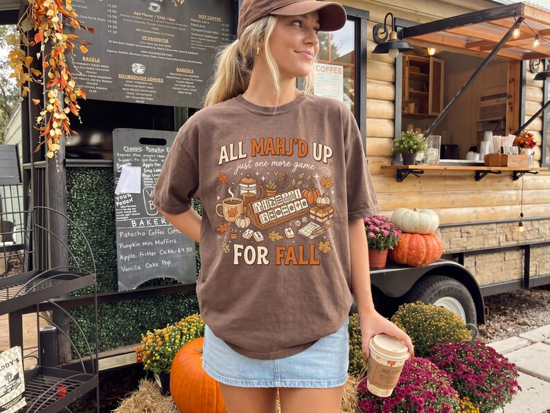 May include: A brown short-sleeve t-shirt featuring a graphic print with the text "ALL MAHJ'D UP just one more game FOR FALL". The design includes illustrations of a mahjong set, a coffee mug, a French press, pumpkins, and autumn leaves.