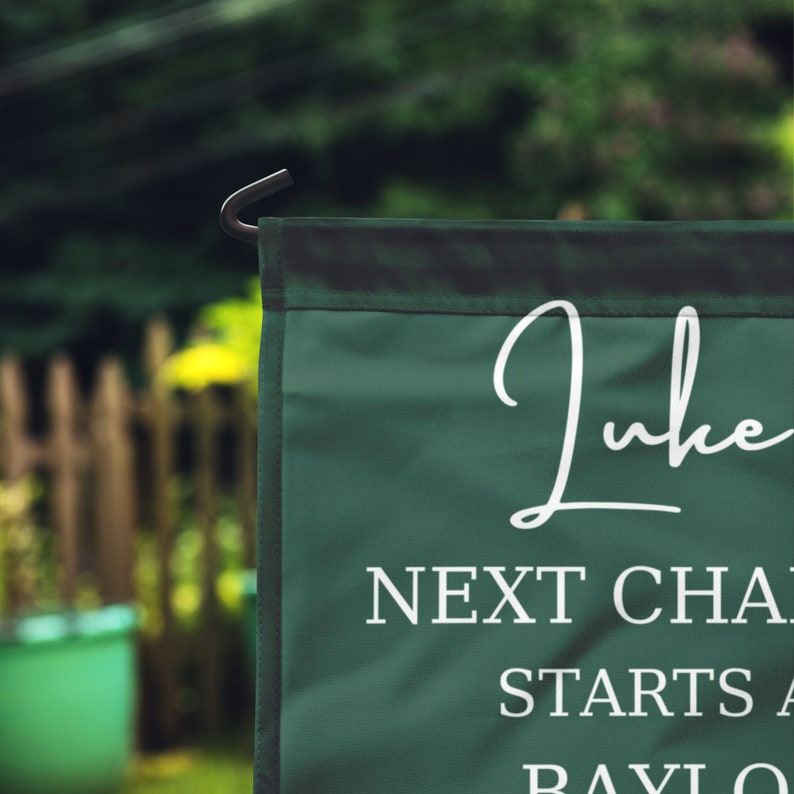 May include: A dark green garden flag with white text. The flag has the name "Luke" in a cursive font, with the words "NEXT CHAPTER STARTS AT BAYLOR" below. The flag is hanging outside in a garden.