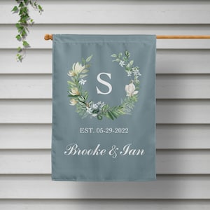 May include: A blue garden flag with a white floral wreath and the letter "S" in the center. The text "EST. 05-29-2022 Brooke & Ian" is printed below the wreath.