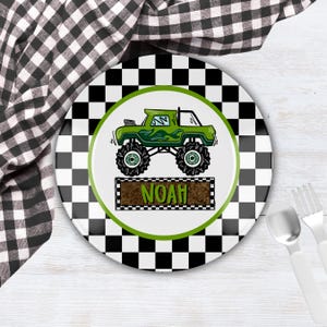 May include: A white ceramic plate with a black and white checkered border. The center features a green monster truck design with the name "NOAH" on a brown rectangular banner. A spoon and fork are visible.