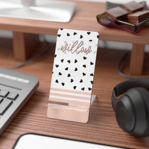 May include: A white phone stand with a black and white heart pattern and the word "willow" in gold lettering. The stand has a pink and white striped design at the bottom and a clear plastic holder for a phone.