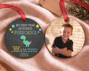 Puede incluir: Dos adornos redondos con cintas rojas. Uno presenta un fondo oscuro con el texto "MY FIRST YEAR OF SCHOOL PRESCHOOL W is for Wesley 2026" y un dinosaurio. El otro muestra una fotografía de un niño sonriente con una camisa negra.