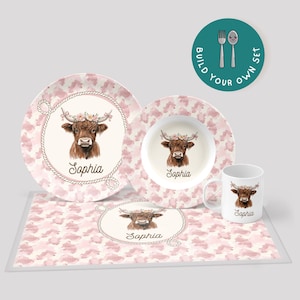 May include: A children's dinnerware set featuring a watercolor illustration of a Highland cow wearing a floral crown. The set includes a plate, bowl, mug, and placemat, all with a pink and white pattern and the name "Sophia".