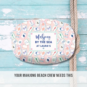 May include: An oval-shaped serving tray with a light pink background, covered in colorful Mahjong tiles. The tray features the text "Mahjong BY THE SEA AT LAURA'S" in a decorative frame. A rope is visible on the right side of the image.