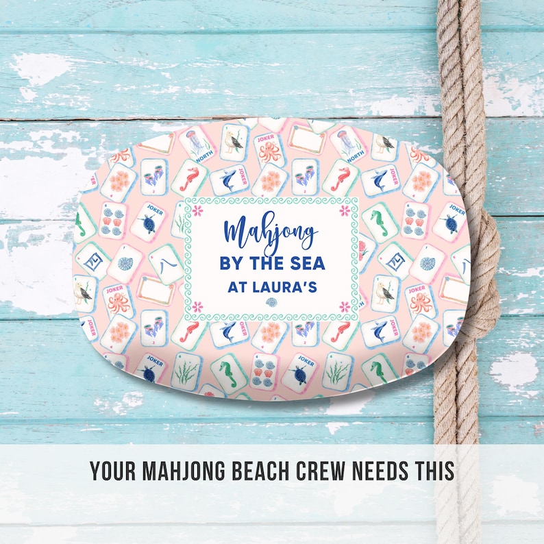 May include: An oval-shaped serving tray with a light pink background, covered in colorful Mahjong tiles. The tray features the text "Mahjong BY THE SEA AT LAURA'S" in a decorative frame. A rope is visible on the right side of the image.