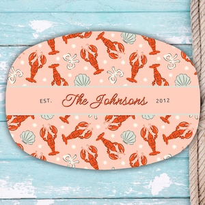 May include: Oval wooden sign with a light coral background featuring a pattern of orange lobsters, white seashells, and floral accents. The sign includes the text "The Johnsons" and "2012". A rope hangs on the right side.