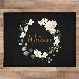 May include: Black doormat with a floral wreath design in white and green. The wreath has gold geometric accents and the word "Welcome" in gold lettering.