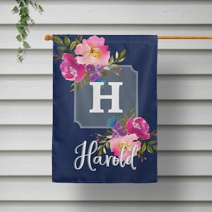 May include: A navy blue garden flag with a white letter H in a gray frame surrounded by pink, purple, and blue watercolor flowers. The text "Harold" is written in a cursive font below the frame.